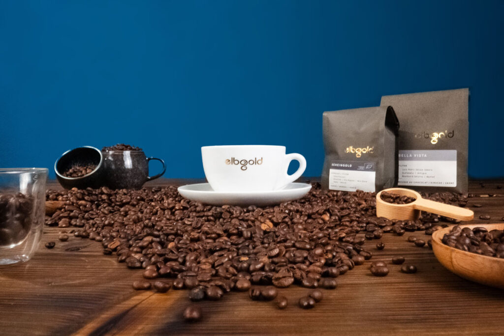 Eine Tasse Elbgold Kaffee auf einer Holzoberfläche umgeben von frischen Kaffeebohnen und einer Packung „Bella Vista“ Filterkaffee.