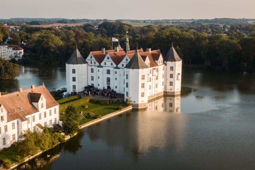 Drohnenaufnahme von Gästen beim 50-jährigen Jubiläum der ttp AG vor Schloss Glücksburg, umgeben von malerischer Natur bei Sonnenuntergang.